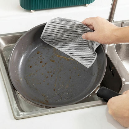 Person cleaning a greasy frying pan with a mesh scrubber in a kitchen sink.